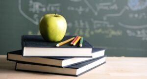 Teacher's desk with apple and pile of books in front of chalkboard