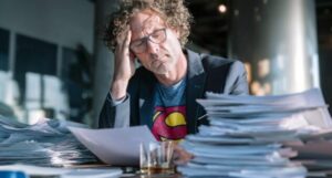 A middle-aged male senior executive sat at a board table. He wears a suit but underneath is a faded superman shirt. He is reading a hard copy of a report and furrowing his brow. He looks tired and has a big pile of other reports to get through. He has a glass of scotch resting on the table.