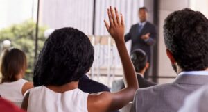 Man taking questions from stage points to woman in audience with raised hand