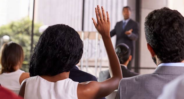 Man taking questions from stage points to woman in audience with raised hand
