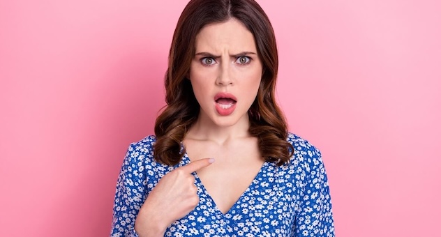 young woman points to herself open-mouthed, looking offended