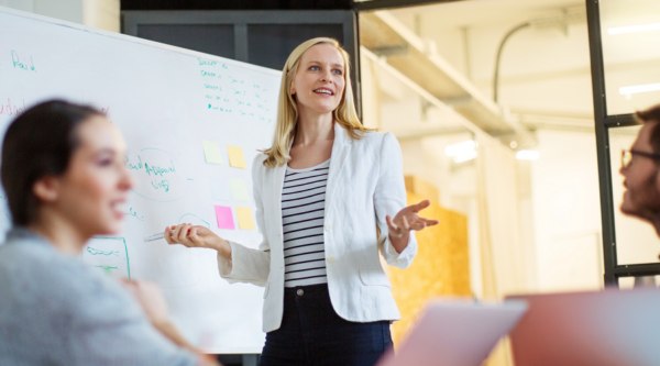 Lady delivering presentation Smiling course leader stands at whiteboard explaining point to engaged group