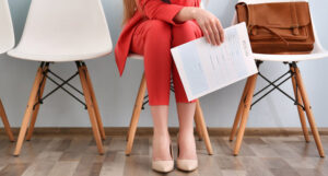 Row of seats with woman in trouser suit in centre seat, resting elbows on her knees and holding a cv