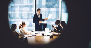 A board meeting with one person standing to present a paper, seen as if through lens