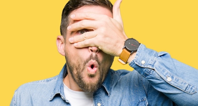 Man with blue eyes and beard covering face and eyes with hand, looking through fingers with scared/embarrassed expression