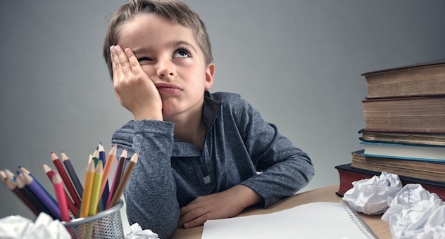 Child with brown hair sits at desk with his head in his hand, looking upwards with unhappy expression. He's surrounded by balled-up paper, a pen pot and a stack of books.