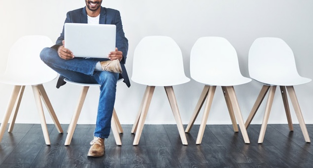 Smiling man on laptop sitting in one of a row of chairs in an empty waiting room