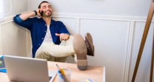 Casually dressed professional leaning back on chair with feet on desk, smiling in relief while talking on the phone