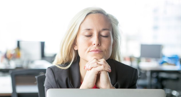 A businesswoman sits at her desk with her eyes closed and her hands clasped under her chin, thinking a decision through.